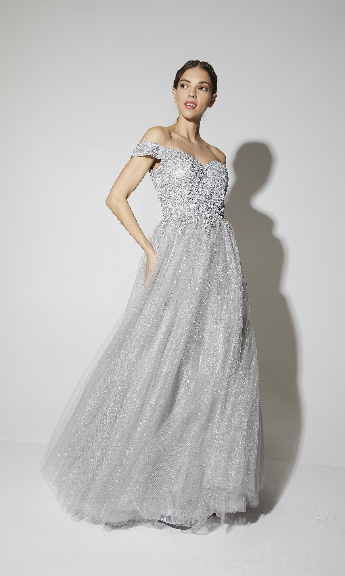 A woman poses in the Off-Shoulder Glitter Prom Ball Gown: Eureka 9899, a light gray dress with a fitted embellished bodice and full glitter-tulle skirt, creating an elegant look against a plain white background.